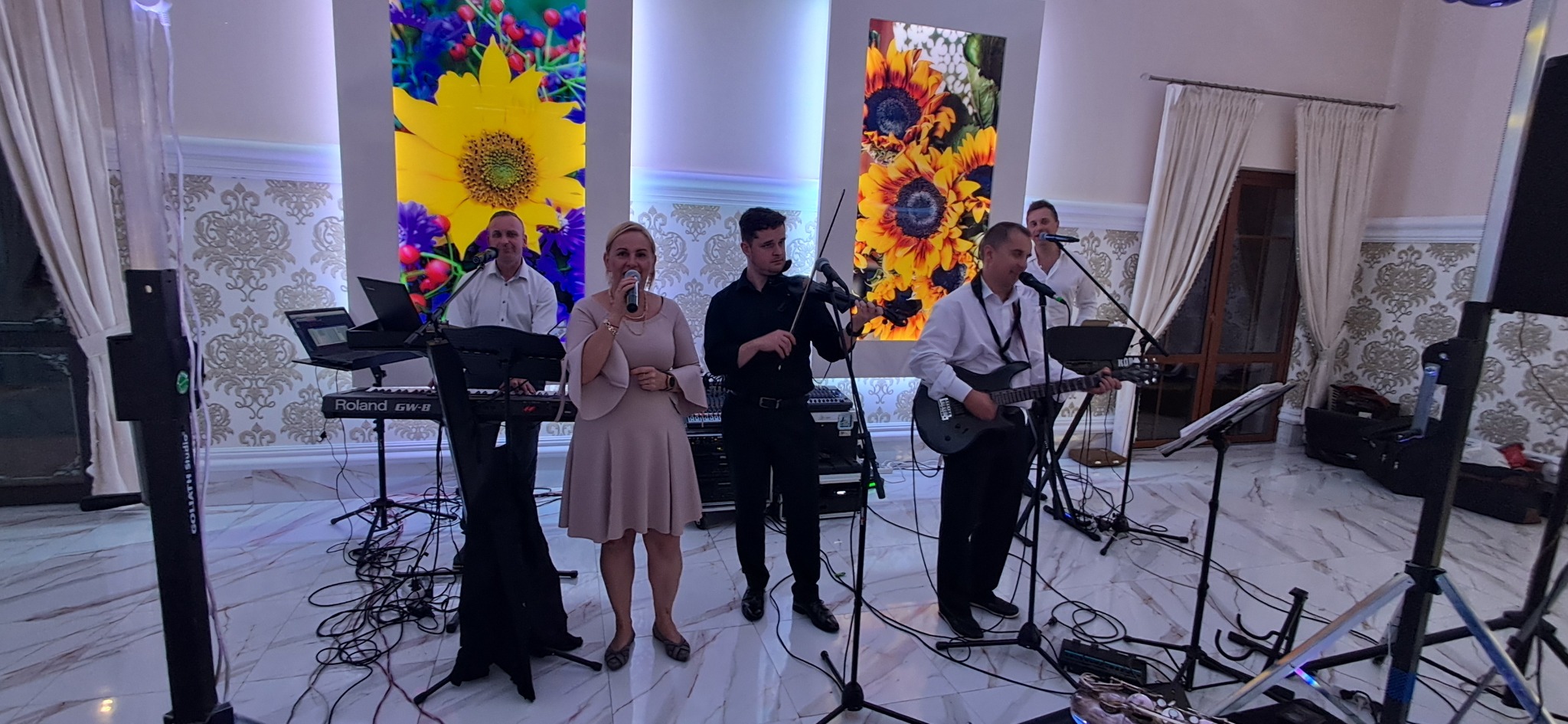 Zespół muzyczny w trakcie występu na sali bankietowej. Wokalistka z mikrofonem, gitarzysta, skrzypek i klawiszowiec. Dekoracje kwiatowe w tle.