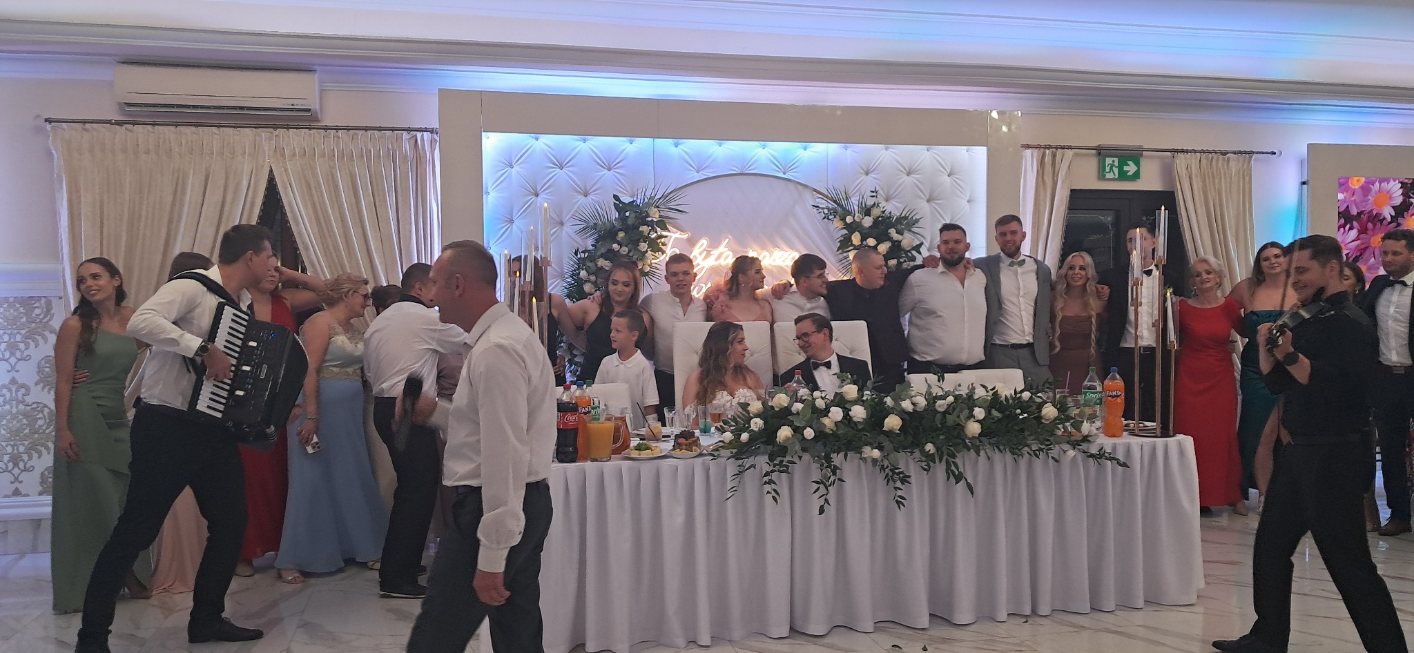 Zespół muzyczny grający na akordeonie podczas wesela, goście weselni pozują do zdjęcia za stołem udekorowanym kwiatami, w tle dekoracja świetlna.