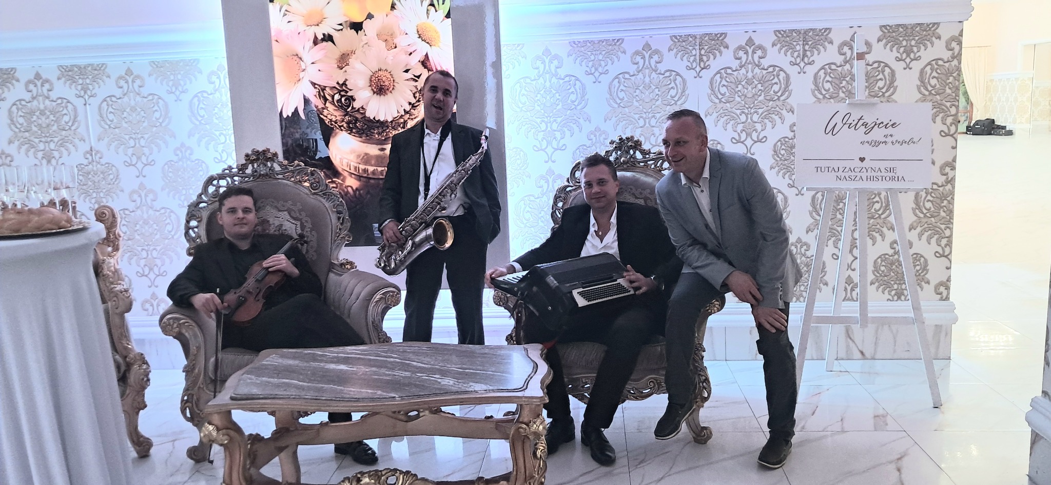 Zespół muzyczny w eleganckich wnętrzach sali weselnej: skrzypek w fotelu, saksofonista, akordeonista i gość weselny. Dekoracje kwiatowe i tablica powitalna.