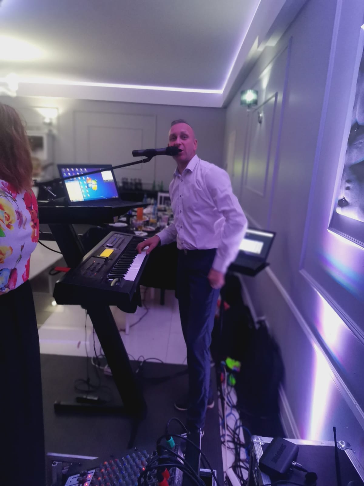 Wokalista zespołu muzycznego w białej koszuli, grający na keyboardzie i śpiewający do mikrofonu podczas występu na tle sali z oświetleniem w odcieniach fioletu.