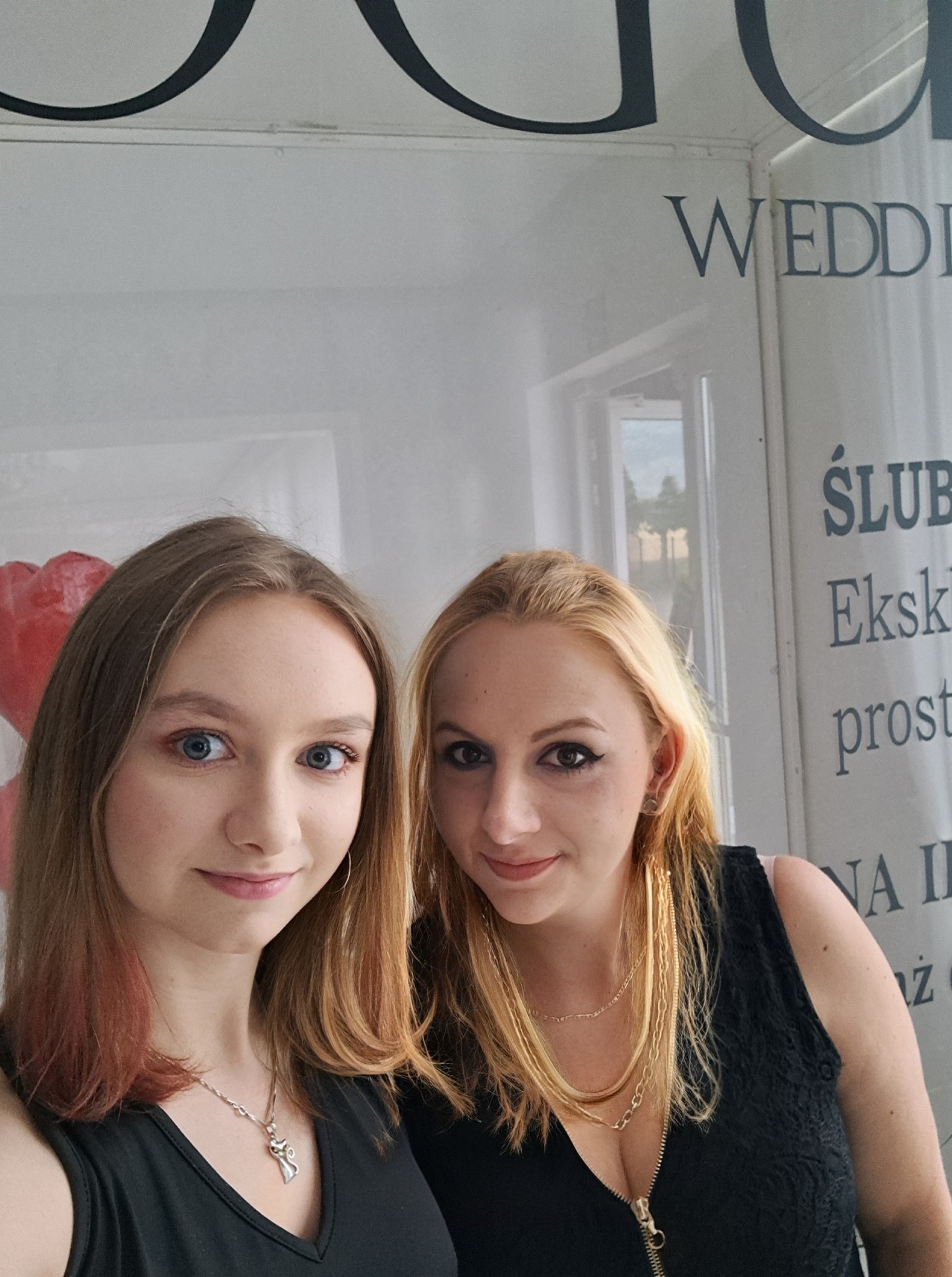 Dwie uśmiechnięte kobiety w czarnych strojach pozują na tle plakatu ślubnego z napisem 'WEDDING'.