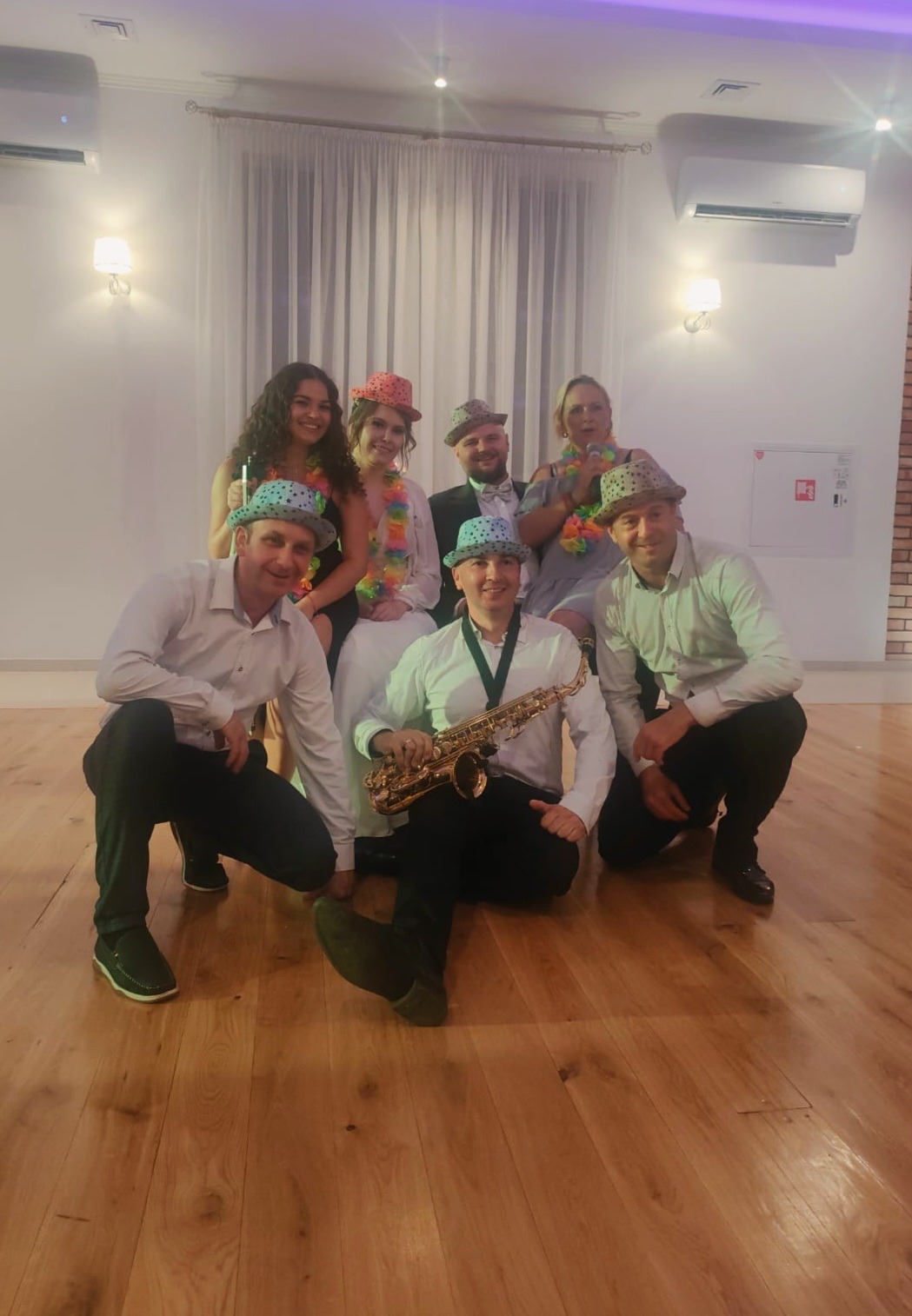 Zespół muzyczny w hawajskich kapeluszach i kwiatach pozuje z saksofonem na parkiecie. W tle białe zasłony i klimatyzator.