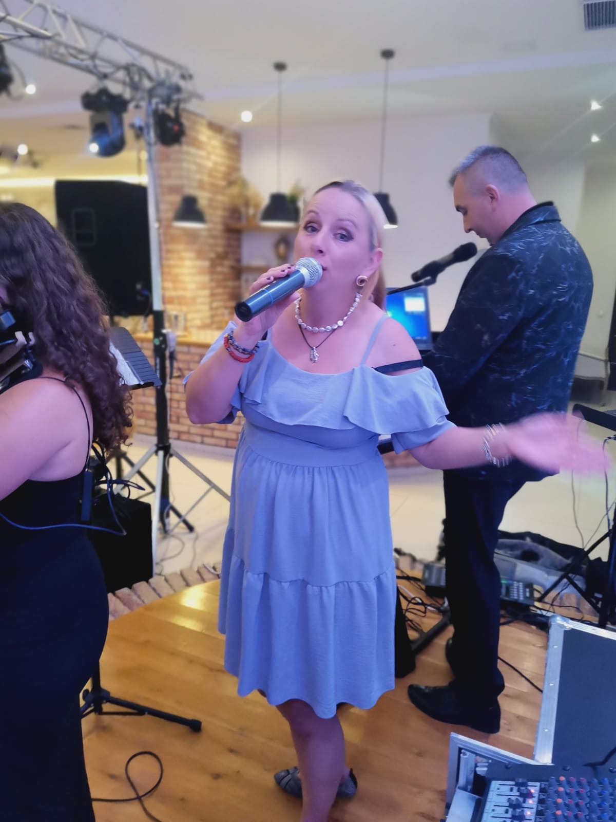 Wokalistka w błękitnej sukience śpiewa do mikrofonu podczas występu zespołu muzycznego na tle ceglanej ściany i oświetlenia scenicznego.