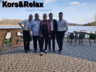 Pięcioosobowy skład muzyczny pozuje na tle jeziora i dekoracyjnych liter, liderka w czarno-czerwonej marynarce.