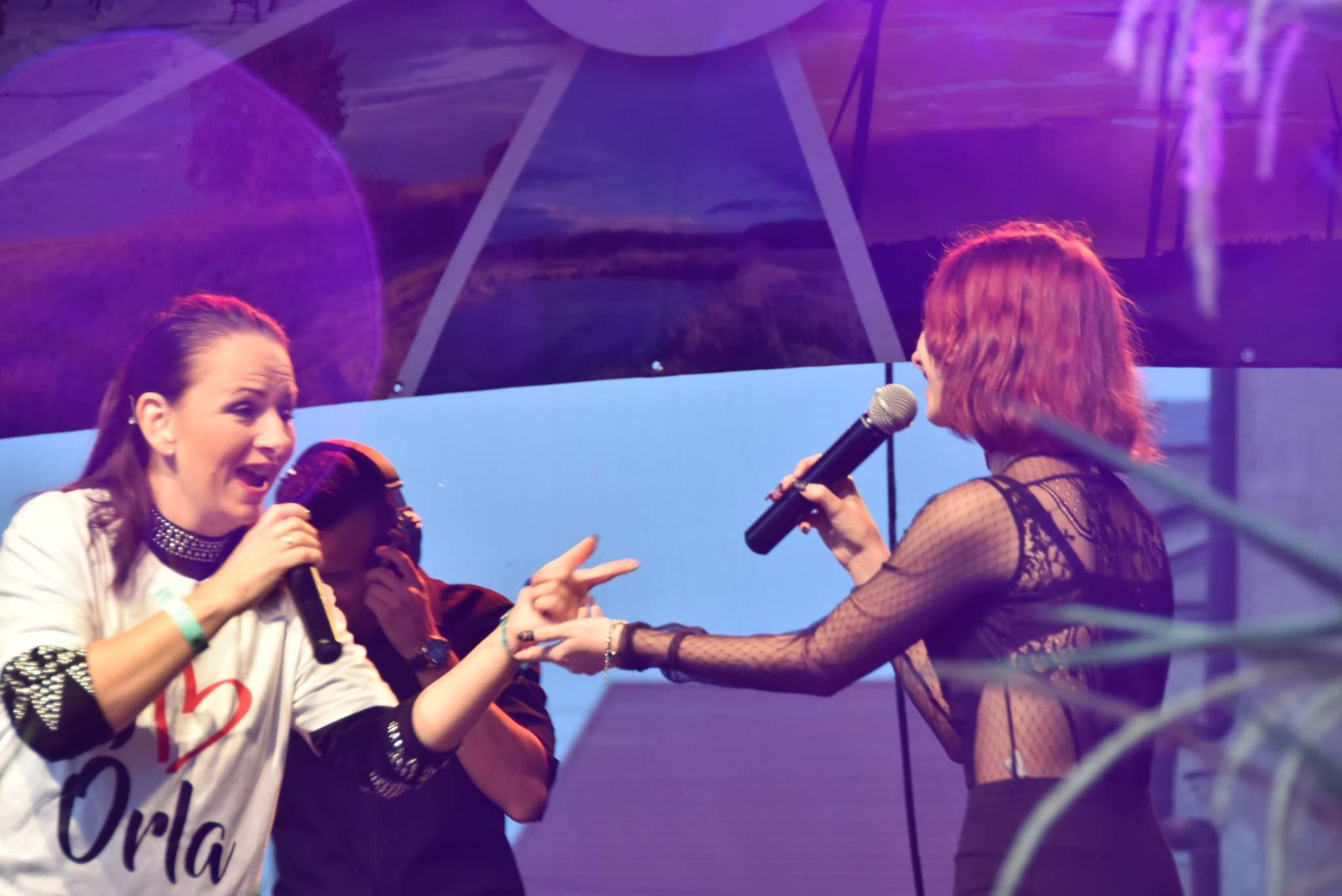 Dwie wokalistki z mikrofonami na scenie podczas występu, jedna w koszulce z napisem 'Orla', druga w bluzce z siateczki, w tle krajobraz z wiatrakami.