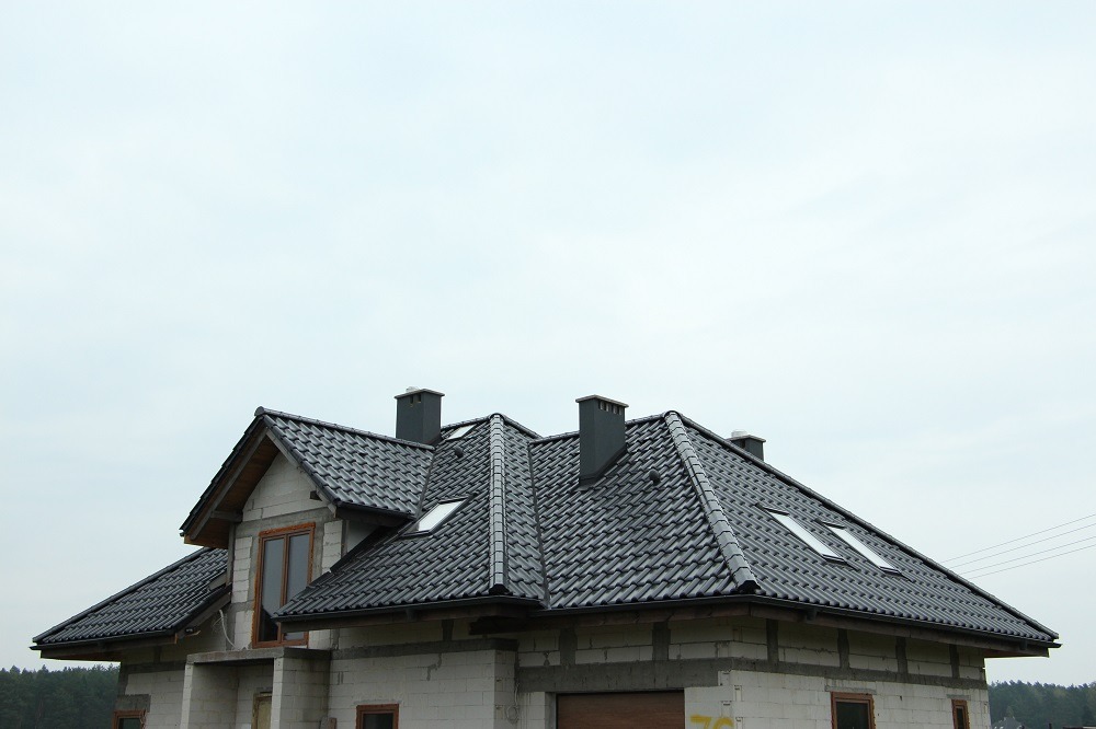 Nowy dom w trakcie budowy z zamontowaną, ciemnoszarą dachówką, oknami dachowymi i kominami wentylacyjnymi, widoczne niezakończone obróbki blacharskie.