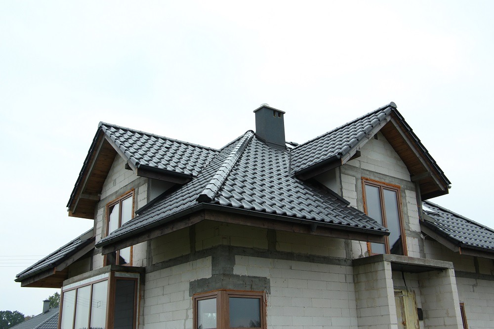 Dom w trakcie budowy z zamontowanym dachem z ciemnej dachówki, widoczne okna i komin, ściany z białego bloczka.