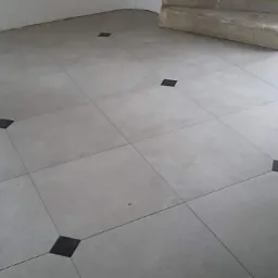 Jasnoszara podłoga z płytek, z czarnymi kwadratami w narożach, w trakcie remontu, widoczne surowe ściany.