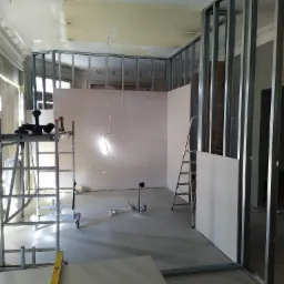 Wnętrze w trakcie remontu z widoczną konstrukcją metalową ścian działowych, częściowo obłożoną płytami gipsowo-kartonowymi, drabiny i narzędzia na podłodze, sufit z pozostałościami po zalaniu.