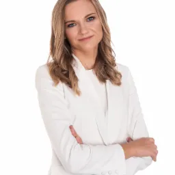 Portret uśmiechniętej kobiety w białym żakiecie, skrzyżowane ręce na jasnym tle.