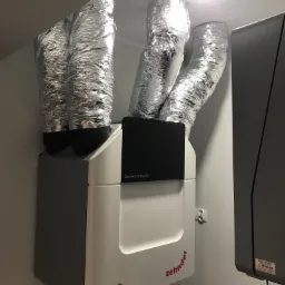 Centrala wentylacyjna Zehnder ComfoAir Q z podłączonymi, izolowanymi przewodami wentylacyjnymi w srebrnej folii, zamontowana przy ścianie w jasnym pomieszczeniu.