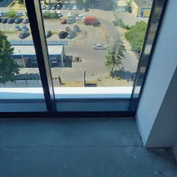 Widok z okna w Gdańsku na parking i budynki w słoneczny dzień, rama okienna z czarnymi profilami, widoczna surowa posadzka.