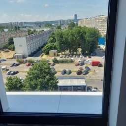 Sebuu - Widok z okna na osiedle mieszkaniowe z parkingiem, drzewami i wieżowcem w oddali, uchwycony w słoneczny dzień.