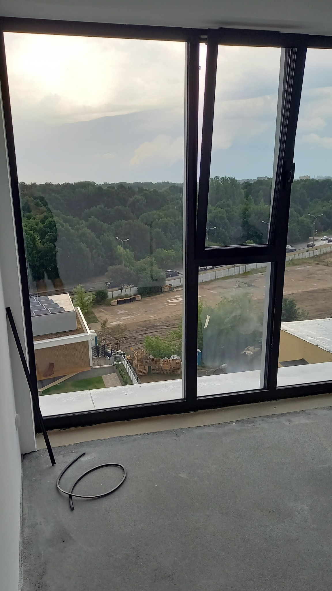 Otwarta okiennica okna z czarną ramą, widok na zewnątrz na budynek z panelami słonecznymi, teren budowy i zieleń, na podłodze leżą elementy uszczelki.