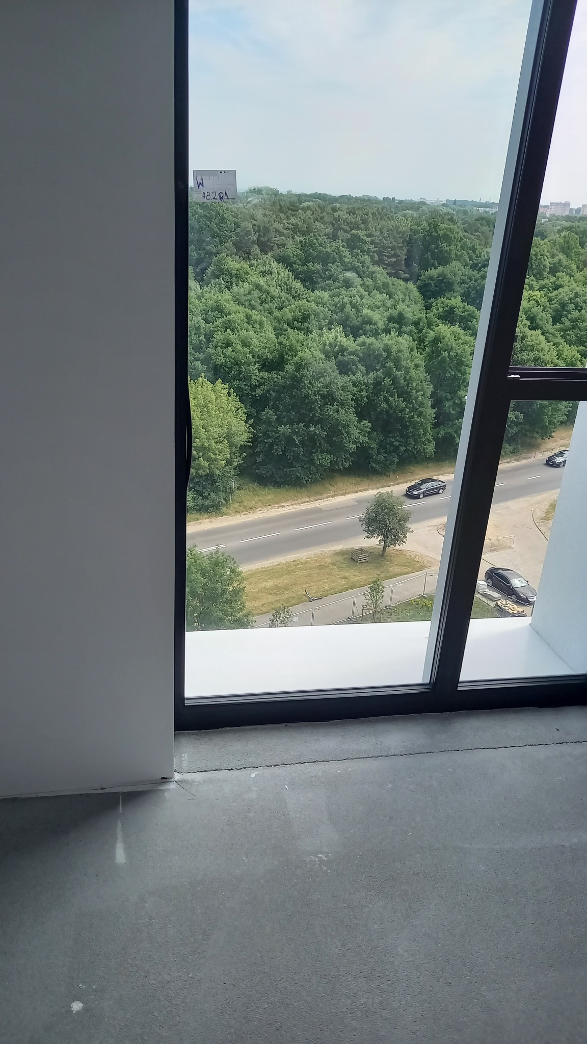 Widok z okna na las i drogę, czarna rama okienna, surowa podłoga, białe ściany