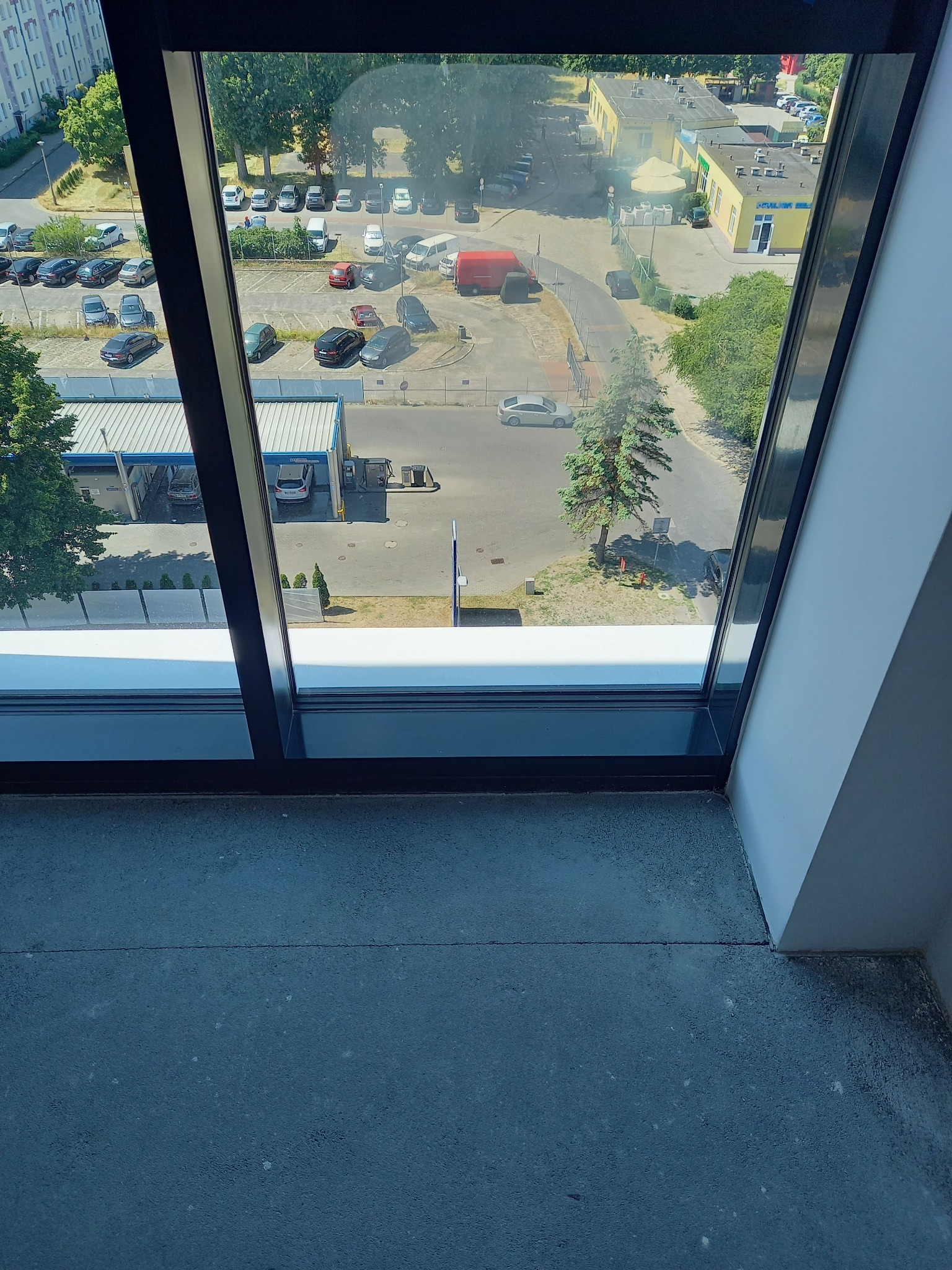 Widok z okna w Gdańsku na parking i budynki w słoneczny dzień, rama okienna z czarnymi profilami, widoczna surowa posadzka.