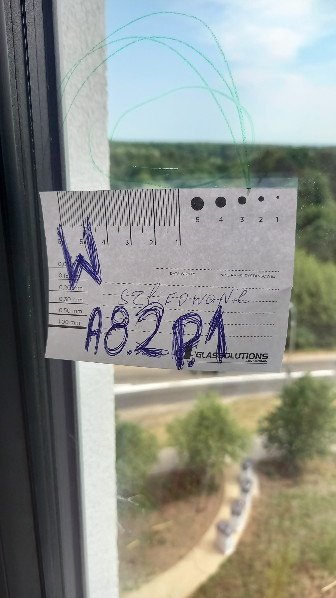 Okna z naklejoną kartką z pomiarami i notatkami 'szlifowane A8.2P1' oraz logo Glassolutions Saint-Gobain, widok na zewnątrz przez szybę z zielonymi bazgrołami.