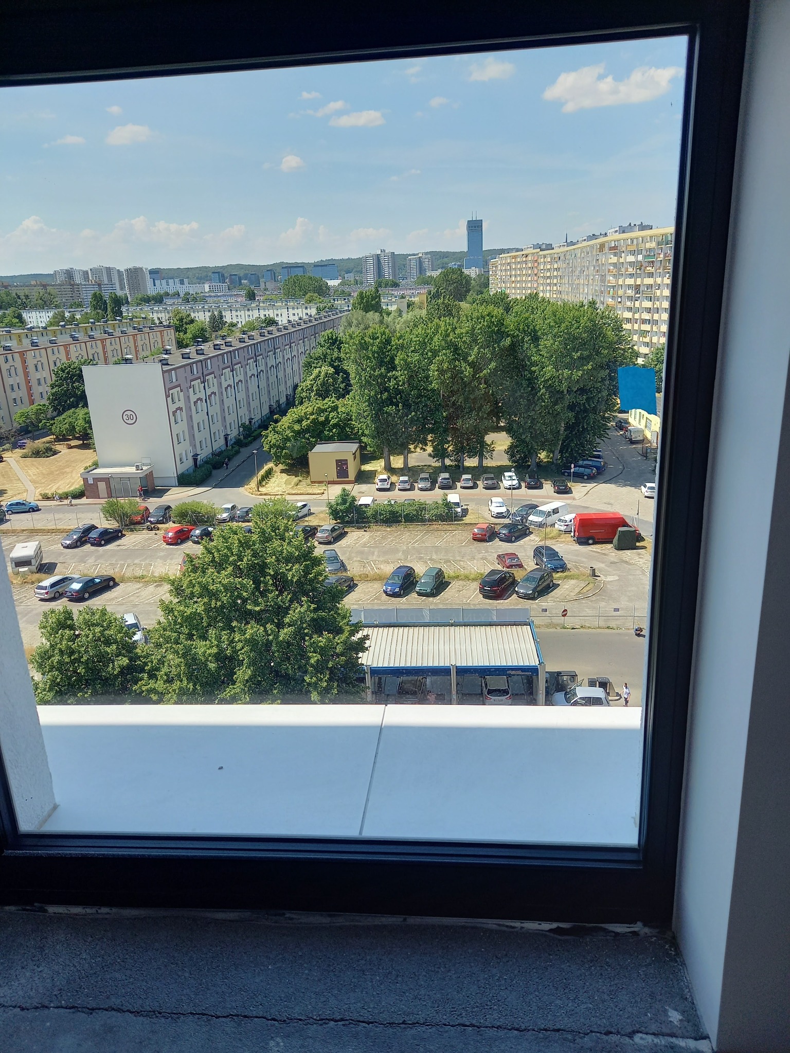 Widok z okna na osiedle mieszkaniowe z parkingiem, drzewami i wieżowcem w oddali, uchwycony w słoneczny dzień.