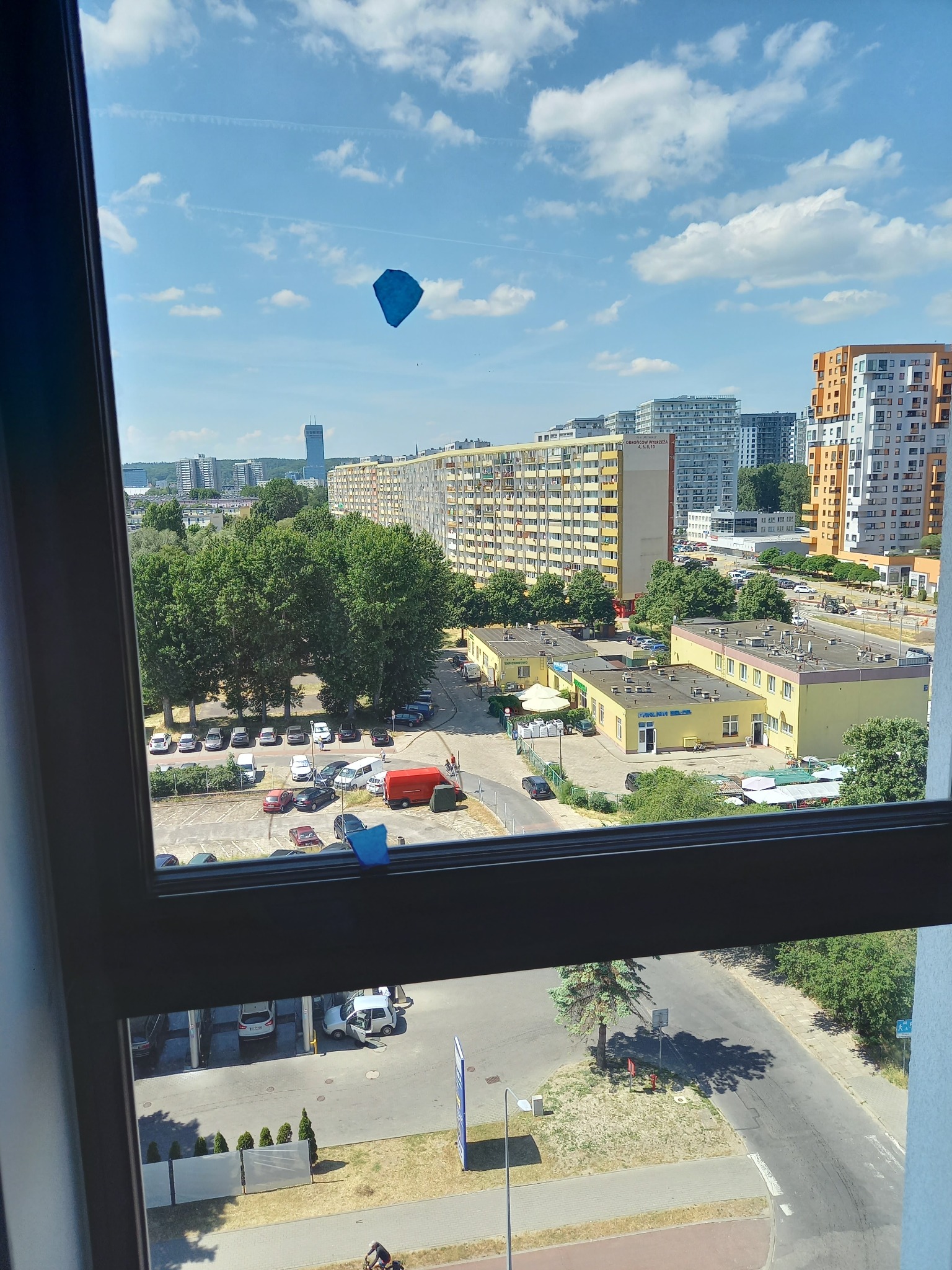 Widok z okna na panoramę miasta z blokami mieszkalnymi, parkingiem i fragmentem ulicy; na szybie okna przyklejone małe, niebieskie karteczki.
