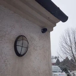 Zewnętrzna kamera monitoringu kopułkowa zamontowana na elewacji budynku obok lampy w stylu marynistycznym, w tle częściowo widoczny samochód dostawczy firmy w zimowej scenerii.