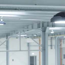 Równomiernie rozmieszczone, przemysłowe lampy LED oświetlają przestronne, jasne wnętrze hali produkcyjnej z widoczną konstrukcją stalową pod sufitem.