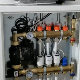 Rozdzielacz ogrzewania podłogowego z przepływomierzami i siłownikami termoelektrycznymi, zamontowany w białej szafce, z widocznym sterownikiem Trinity na górze.