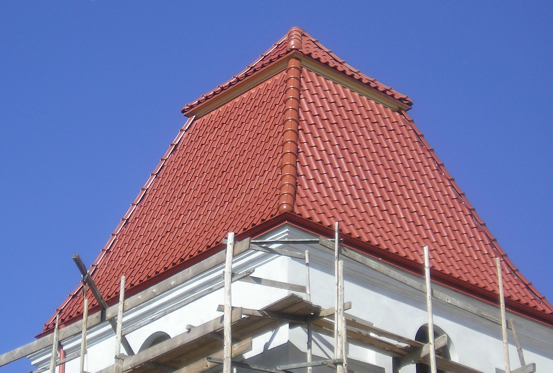 Wieża kościoła z nową, czerwoną dachówką karpiówką, widoczne rusztowanie i obróbki blacharskie, błękitne niebo w tle.