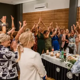 Rozentuzjazmowane kobiety w różnym wieku, ubrane w eleganckie stroje, z podniesionymi rękami i uśmiechami, świętują przy stole udekorowanym świecami i napojami podczas wydarzenia w sali z drewnianą...