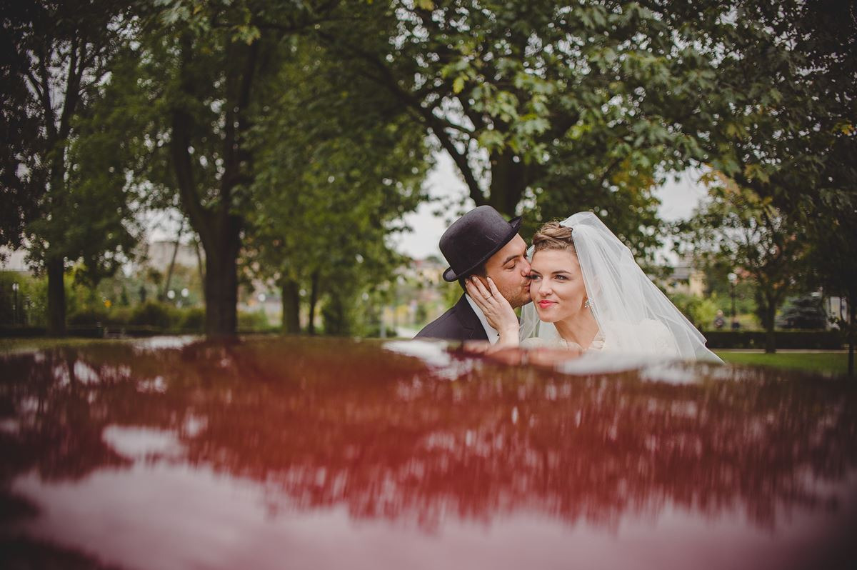 Para młoda: pan młody w czarnym meloniku całuje pannę młodą w welonie na tle parku, odbicie w lakierowanej powierzchni.
