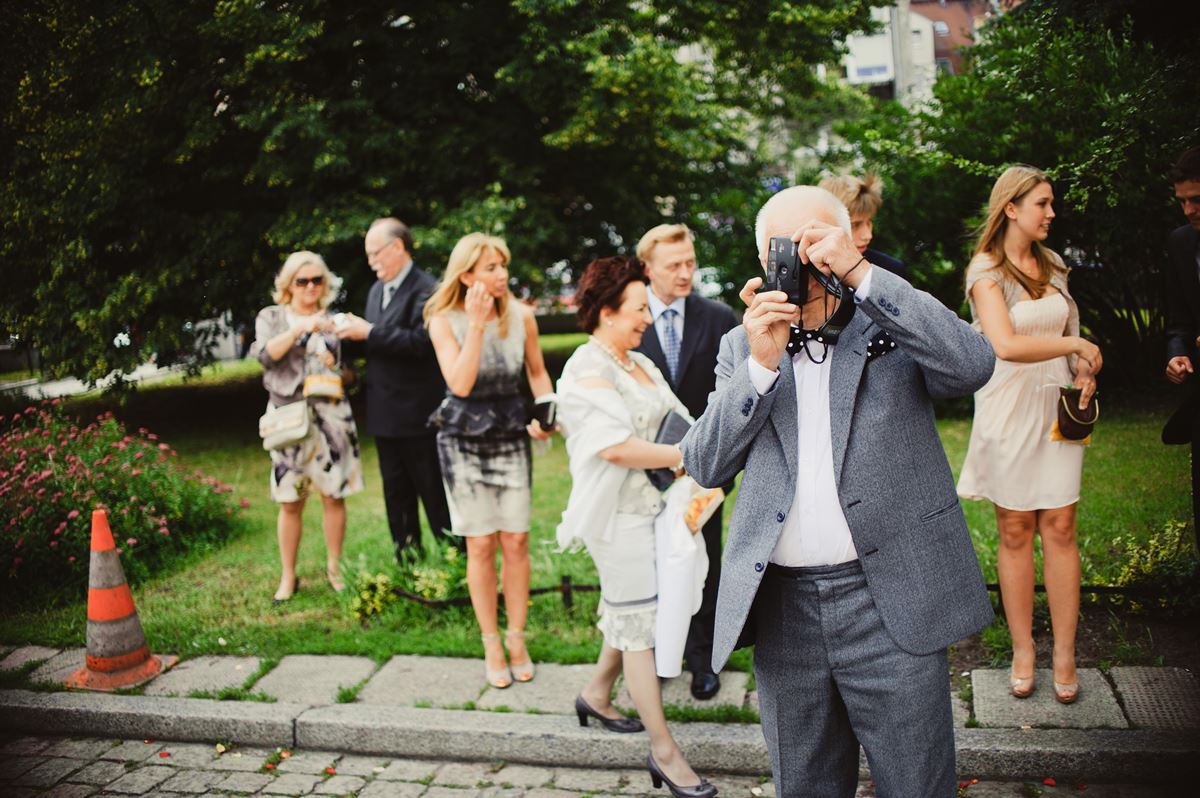 Mężczyzna w szarym garniturze i muszce w kropki robi zdjęcie grupie osób na zewnątrz, używając aparatu retro.
