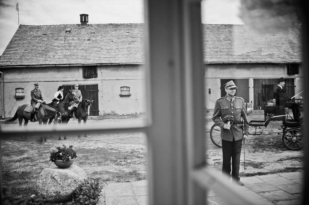 Czarno-białe zdjęcie przez okno: mężczyzna w mundurze z bukietem, w tle konie i bryczka na tle starego budynku.