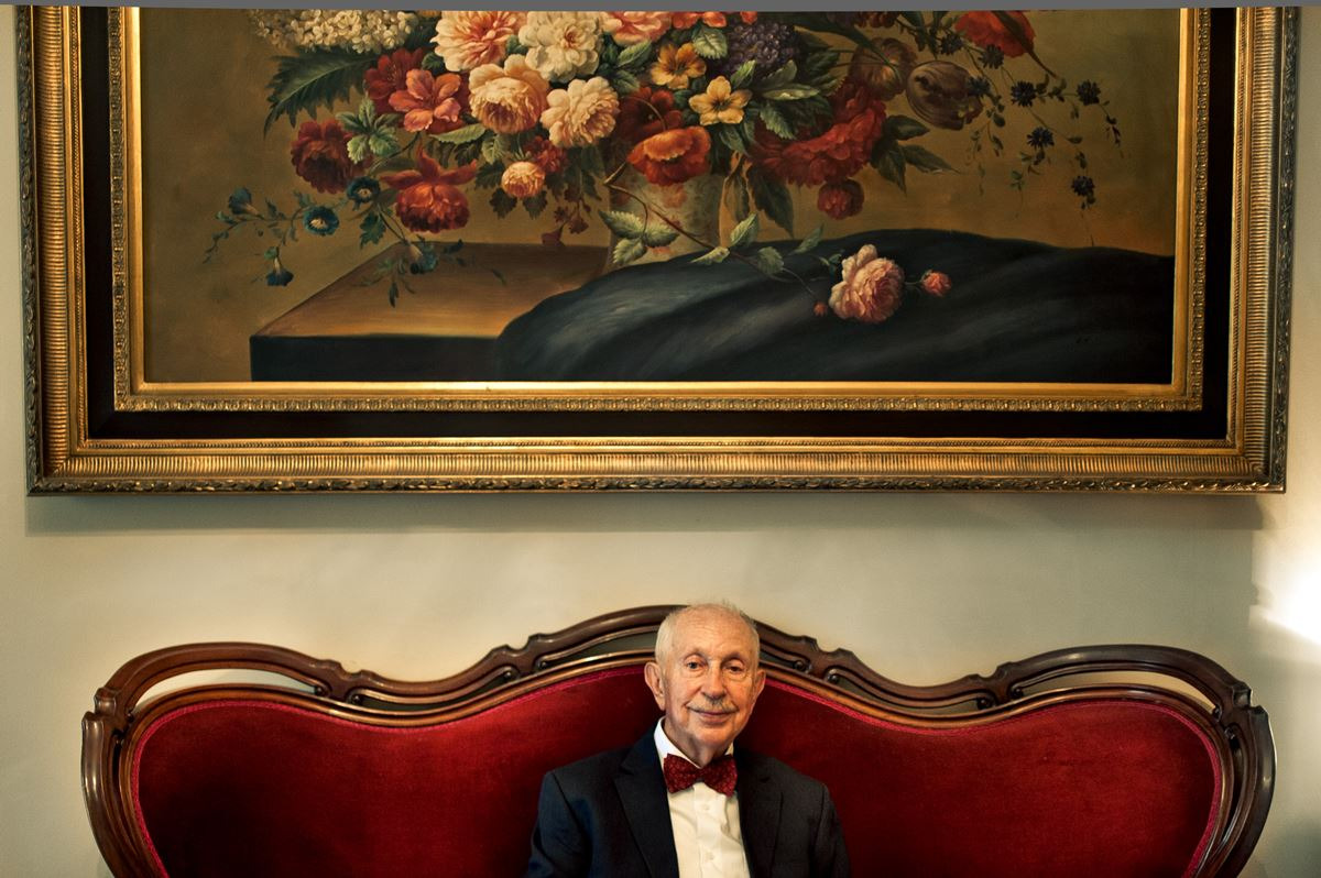Starszy mężczyzna w garniturze i muszce siedzi na czerwonej, aksamitnej sofie pod obrazem martwej natury w złotej ramie.