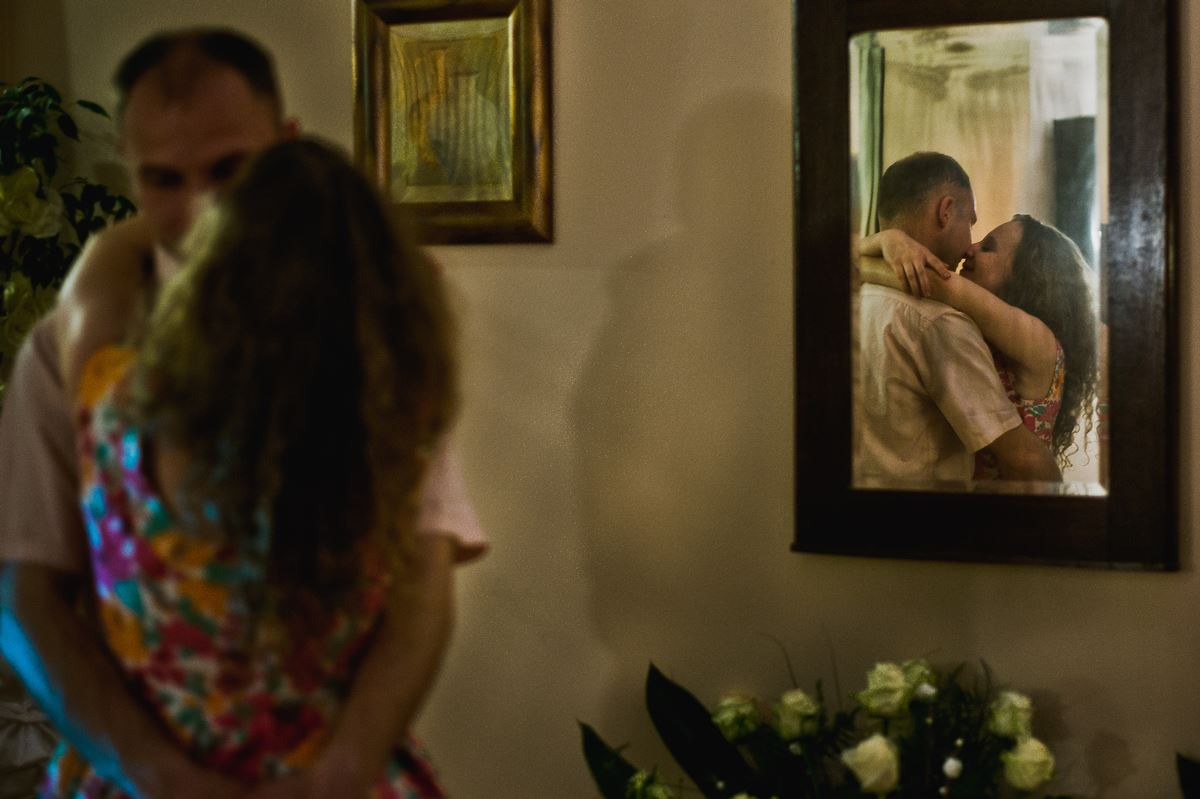 Para obejmująca się, widoczna w lustrze na ścianie, z bukietem białych kwiatów w rogu. Kobieta w kwiecistej sukience, mężczyzna w jasnej koszuli.