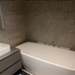 Nowoczesna łazienka z białą wanną wolnostojącą i umywalką nablatową na szarej szafce, ściany wyłożone mozaiką z płytek w odcieniach szarości.