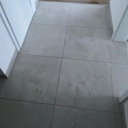 FORADEA SPÓŁKA Z OGRANICZONĄ ODPOWIEDZIALNOŚCIĄ - Ułożone szare płytki podłogowe w przedpokoju, widoczne fragmenty białych drzwi i drewnianej podłogi. Minimalistyczne wykończenie wnętrza.