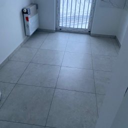 FORADEA SPÓŁKA Z OGRANICZONĄ ODPOWIEDZIALNOŚCIĄ - Wykończone, szare płytki podłogowe w jasnym pomieszczeniu z grzejnikiem w folii i oknem balkonowym z widokiem na zaśnieżone pole.