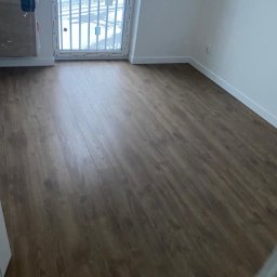 FORADEA SPÓŁKA Z OGRANICZONĄ ODPOWIEDZIALNOŚCIĄ - Ułożone panele podłogowe w pokoju z oknem w Warszawie. Widoczna faktura drewna i białe listwy przypodłogowe, grzejnik. Świeżo po remoncie.