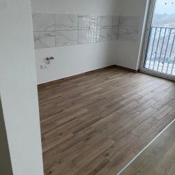 FORADEA SPÓŁKA Z OGRANICZONĄ ODPOWIEDZIALNOŚCIĄ - Wykończona kuchnia w nowym budownictwie: brązowe panele podłogowe imitujące drewno, białe ściany z marmurową glazurą i widok na zewnątrz przez okno balkonowe.