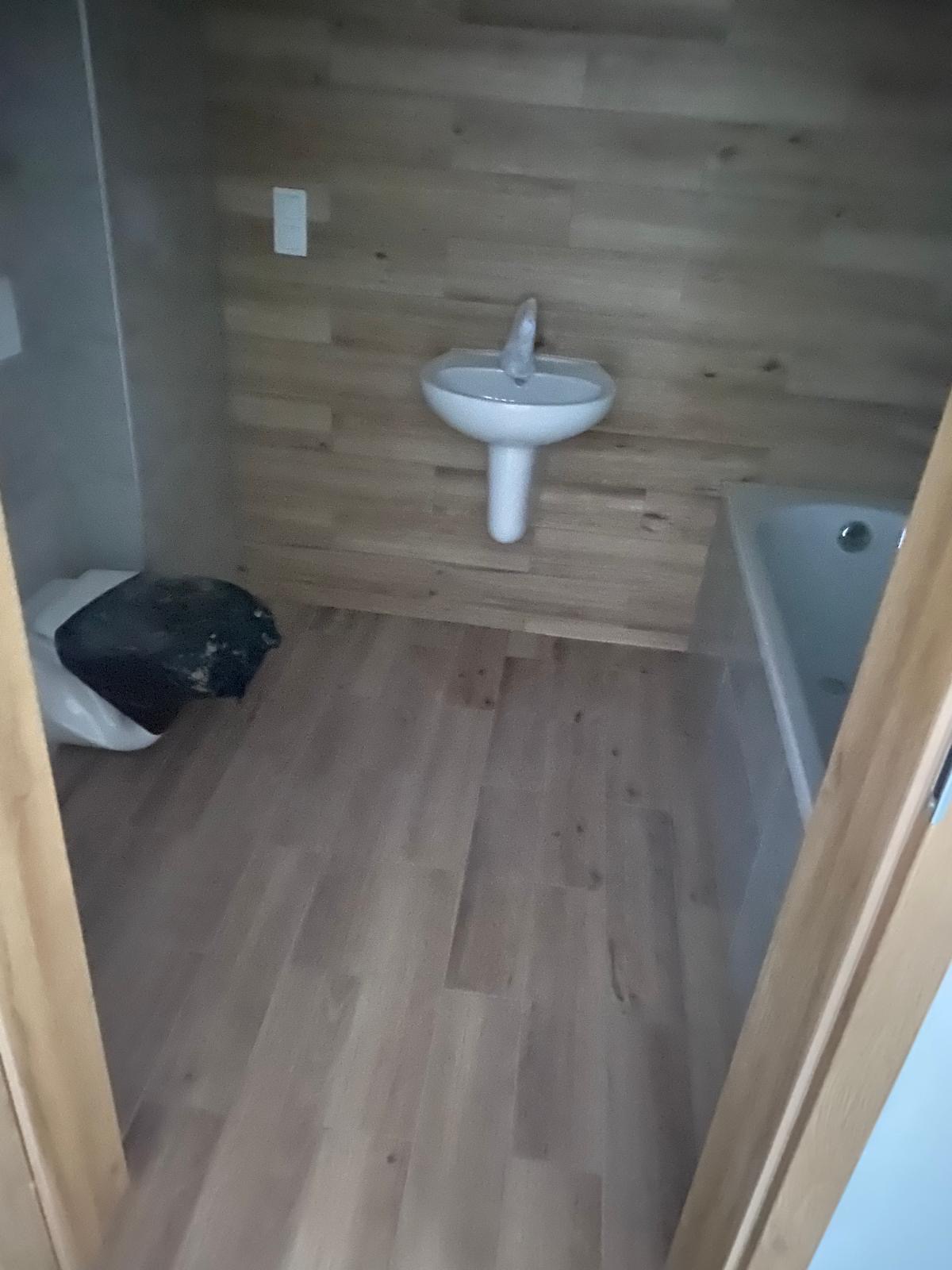 Wykończona łazienka z drewnianą okładziną ścienną i podłogą, białą umywalką na postumencie i wanną. Widoczny worek z odpadami.