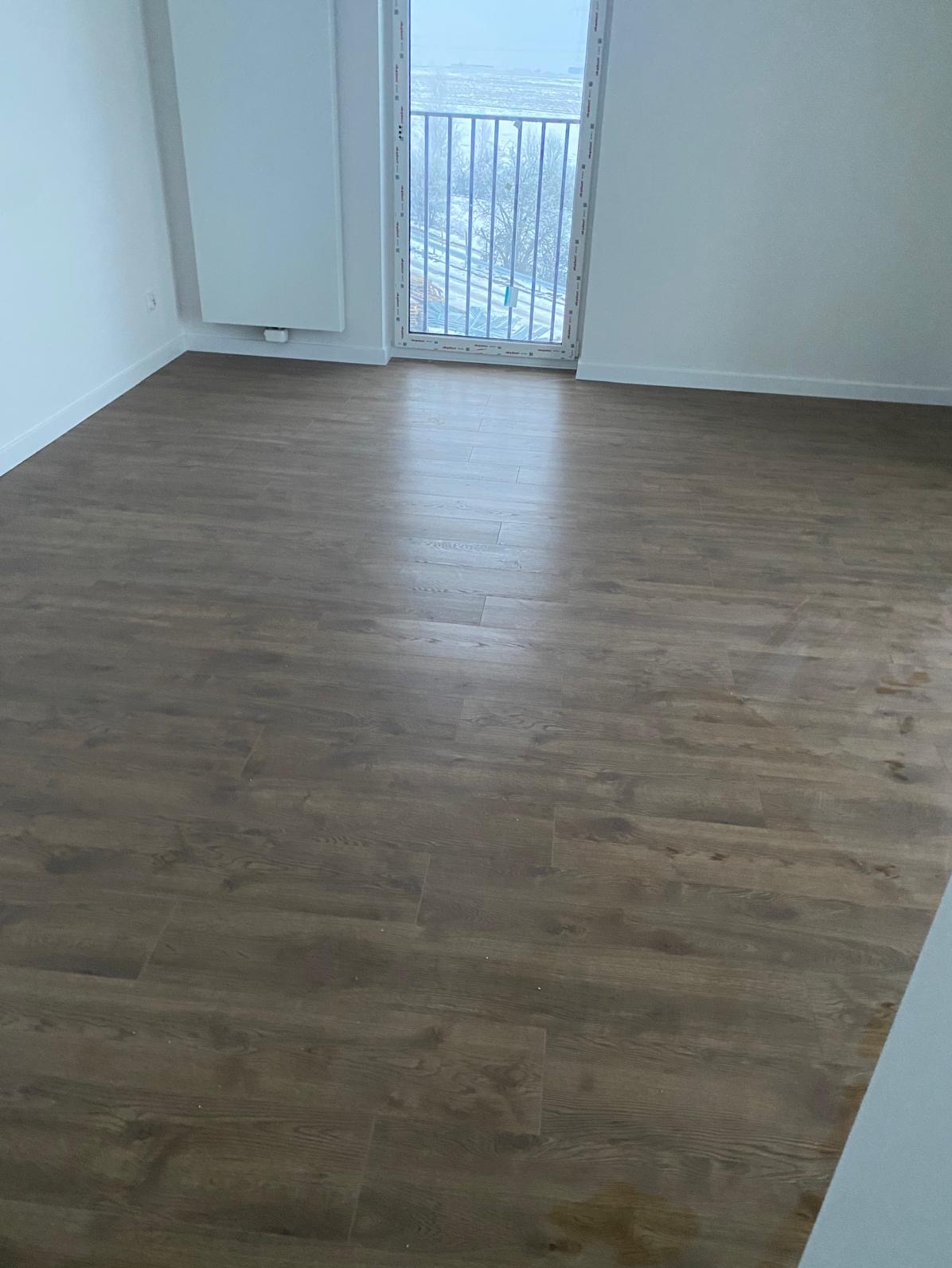 Nowe, ciemne panele podłogowe w pustym pokoju z białymi ścianami i balkonem. Widoczne detale faktury drewna i równo ułożone deski. Minimalistyczne wnętrze.