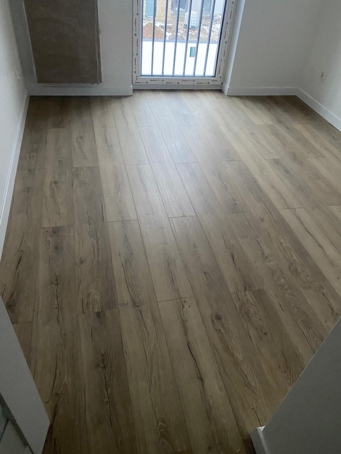 Jasne panele podłogowe z widoczną strukturą drewna w nowym pokoju z oknem balkonowym. Wykończenie w neutralnych odcieniach bieli i beżu. Minimalistyczny styl.