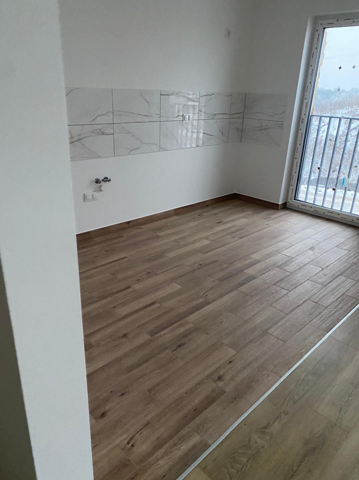 Wykończona kuchnia w nowym budownictwie: brązowe panele podłogowe imitujące drewno, białe ściany z marmurową glazurą i widok na zewnątrz przez okno balkonowe.