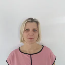 Portret kobiety o blond włosach i niebieskich oczach, ubranej w różową bluzkę z czarną lamówką, pozującej na białym tle.