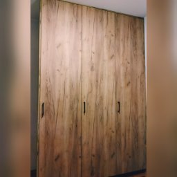 PRW Chud-Meb - Wysoka, trzydrzwiowa szafa z frontami imitującymi naturalne drewno o jasnobrązowym odcieniu i wyraźnym usłojeniu, wyposażona w minimalistyczne, pionowe, czarne uchwyty.