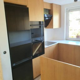 PRW Chud-Meb - Jasna kuchnia z drewnianymi frontami, czarnym piekarnikiem i lodówką w zabudowie, biały blat roboczy przy oknie.