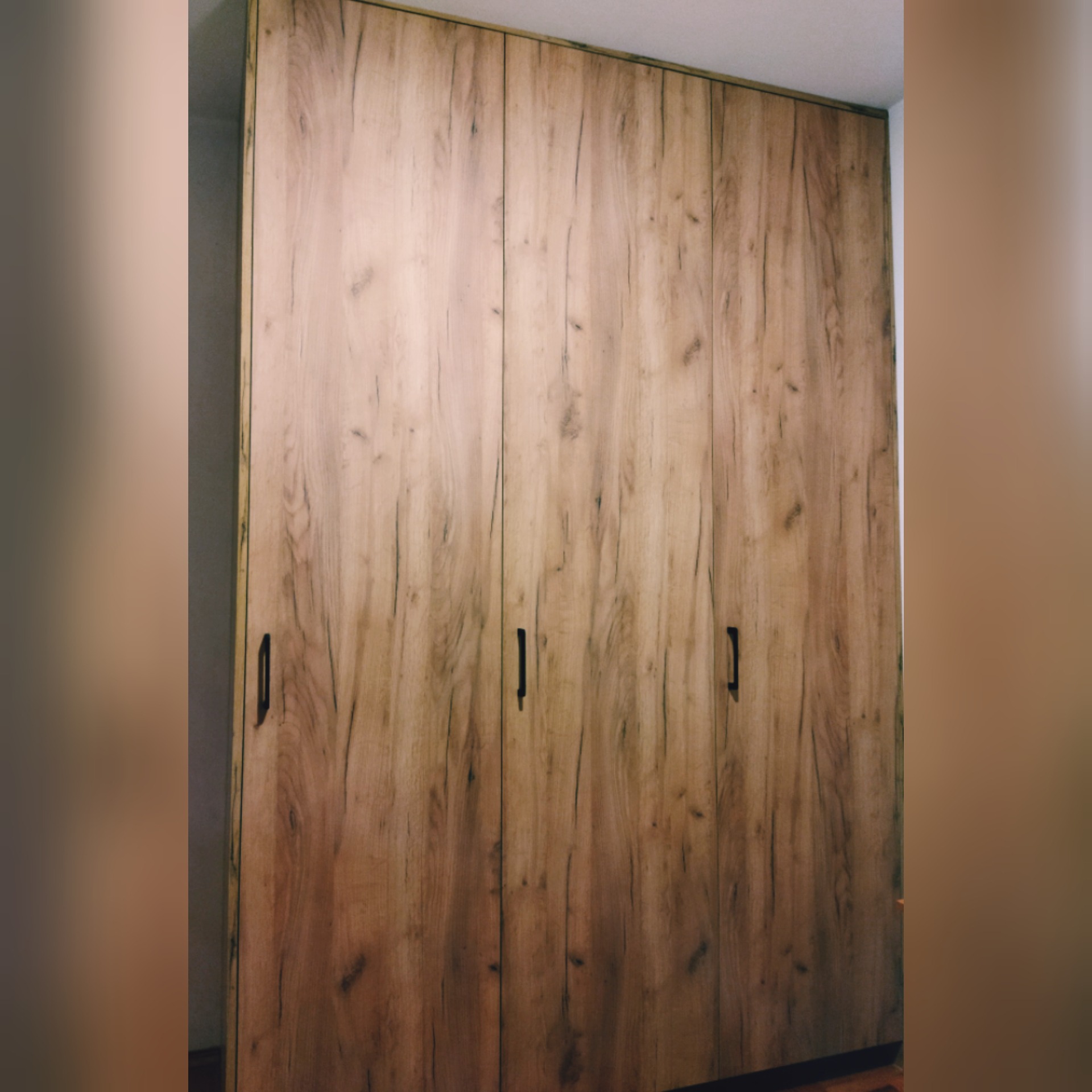 Wysoka, trzydrzwiowa szafa z frontami imitującymi naturalne drewno o jasnobrązowym odcieniu i wyraźnym usłojeniu, wyposażona w minimalistyczne, pionowe, czarne uchwyty.