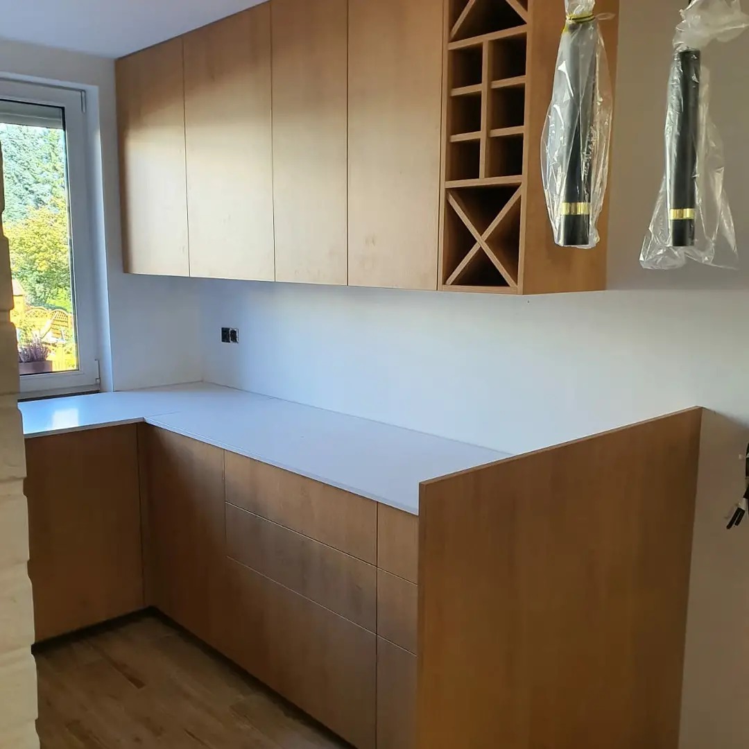 Kuchnia na wymiar z drewnianymi frontami, białym blatem i otwartą półką na wino, widoczne lampy w folii ochronnej.