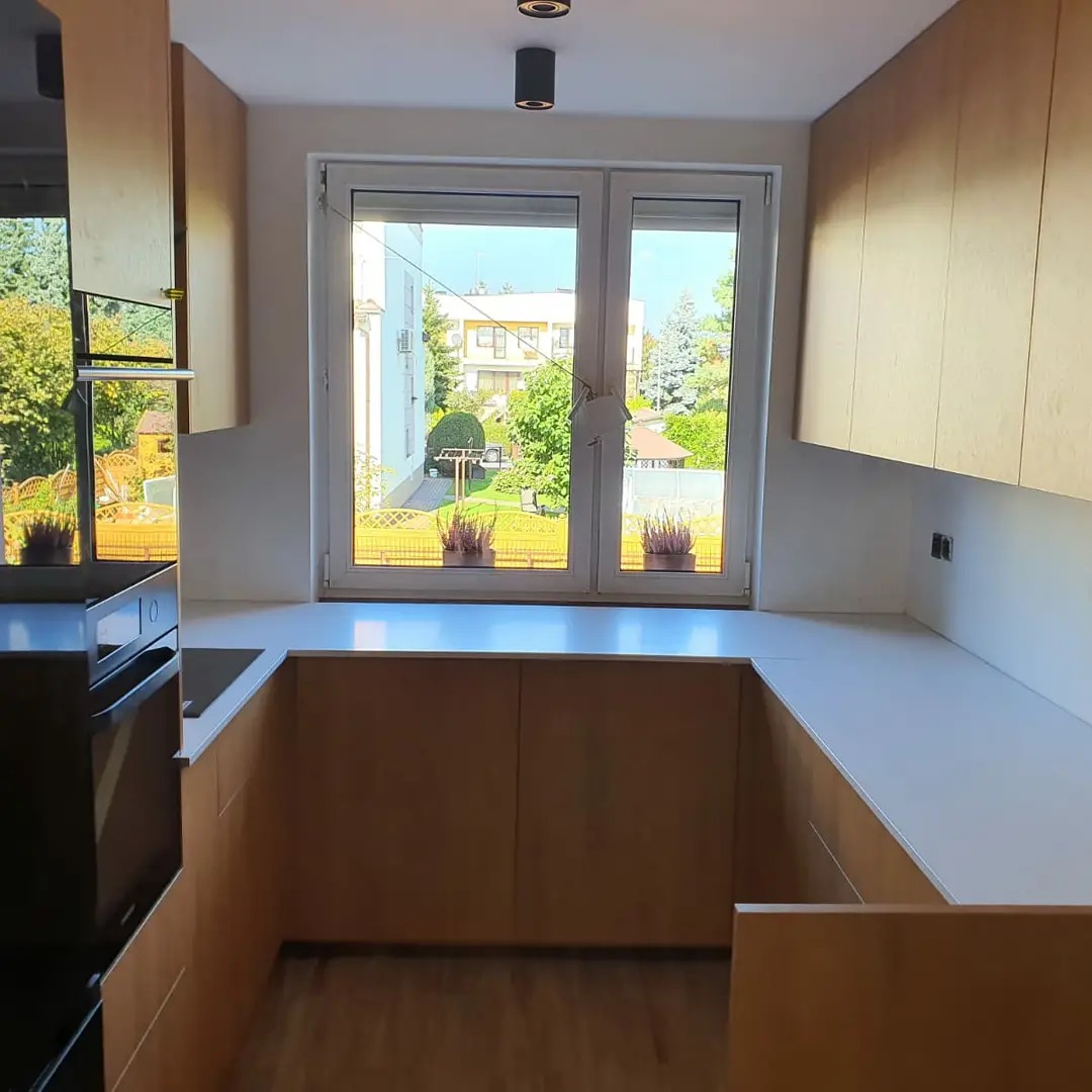 Nowoczesna kuchnia z jasnymi drewnianymi frontami, białym blatem i widokiem na ogród przez okno.