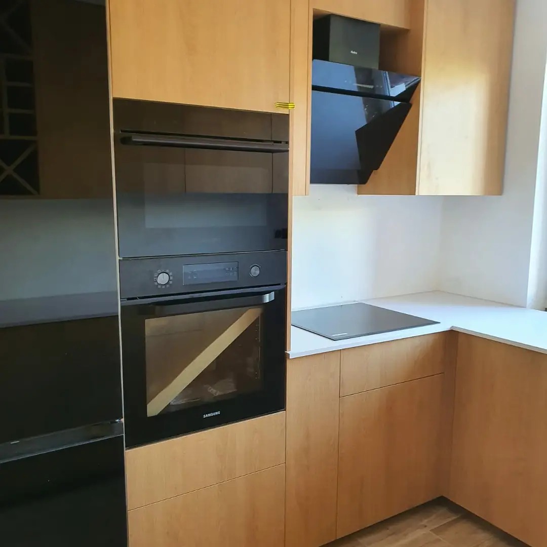 Fragment zabudowy kuchennej z drewnianymi frontami, piekarnik Samsung w zabudowie, płyta indukcyjna i czarny okap podszafkowy.