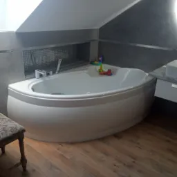 Nowoczesna, narożna wanna w łazience na poddaszu, wykończona szarą mozaiką i drewnopodobnymi płytkami podłogowymi, z dziecięcymi zabawkami na brzegu wanny.